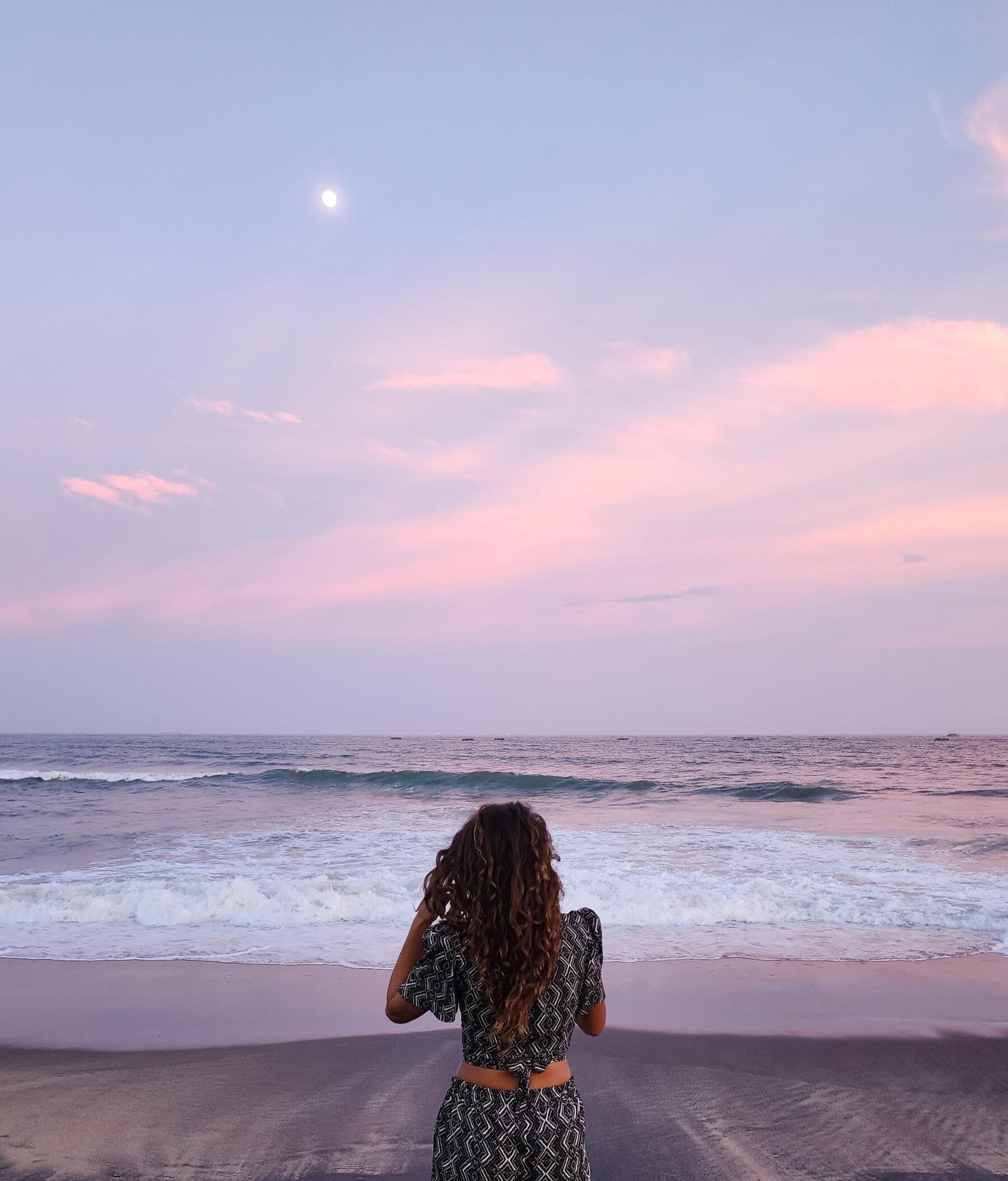 Samara standing by the ocean at sunset, facing the waves under a soft pink and purple sky.