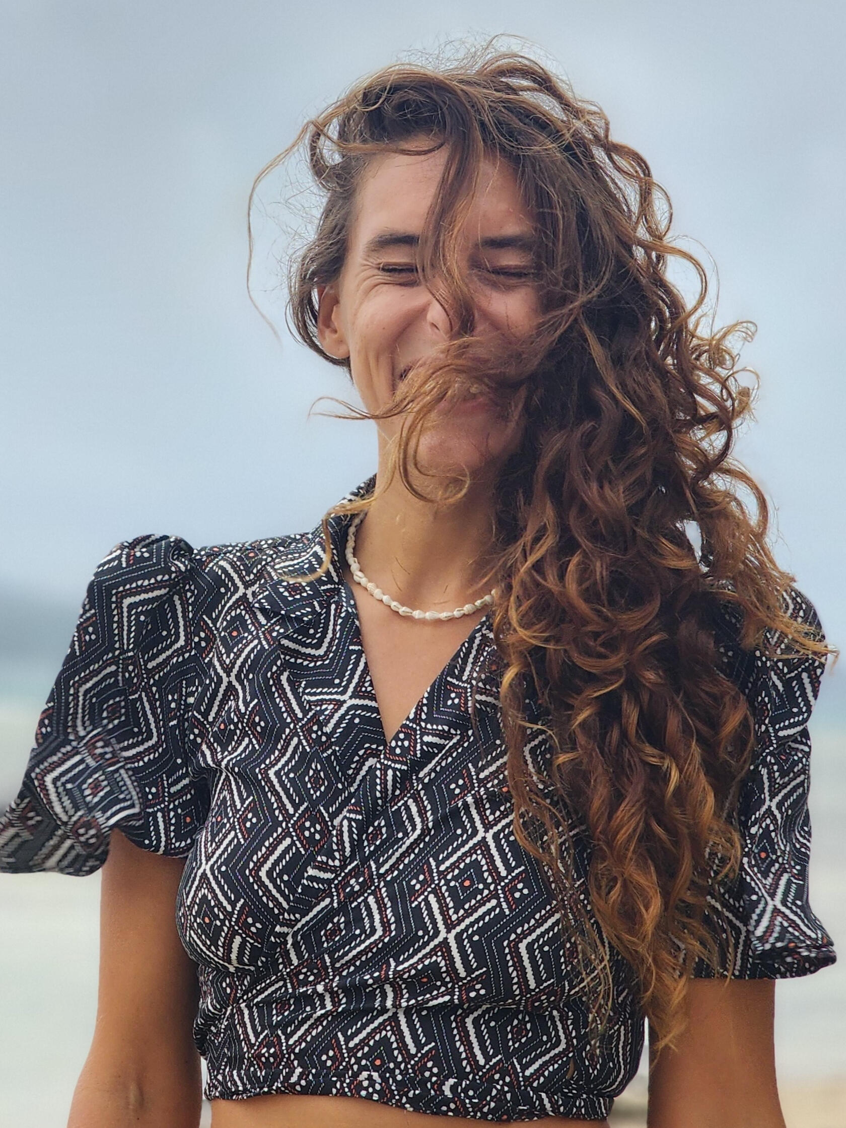 Samara Ruighaver, Founder of Inner Shift Wellness Coaching Samara Ruighaver smiling by the ocean with curly hair blowing in the wind, creating a relaxed and grounded feeling.